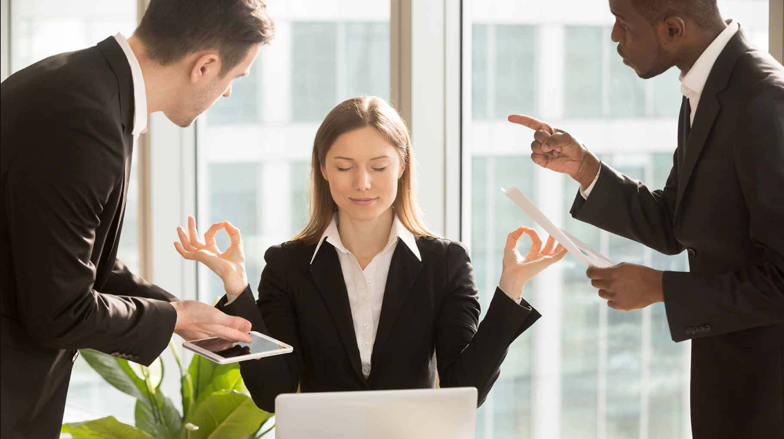 Uma mulher em um ambiente de escritório, praticando meditação para manter a calma, enquanto dois colegas discutem ao seu redor. A imagem reflete a importância de manter a calma e o equilíbrio no trabalho, especialmente para quem lida com o gerenciamento de Social Media, onde as situações podem ser tensas e exigem controle emocional.