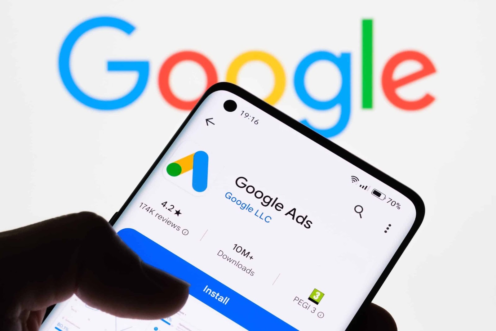 Uma mão segura um smartphone com o aplicativo Google Ads aberto na tela, mostrando a interface de download e avaliação do app, com o logotipo do Google ao fundo. A imagem ilustra a gestão de campanhas de marketing digital e publicidade paga na plataforma Google.