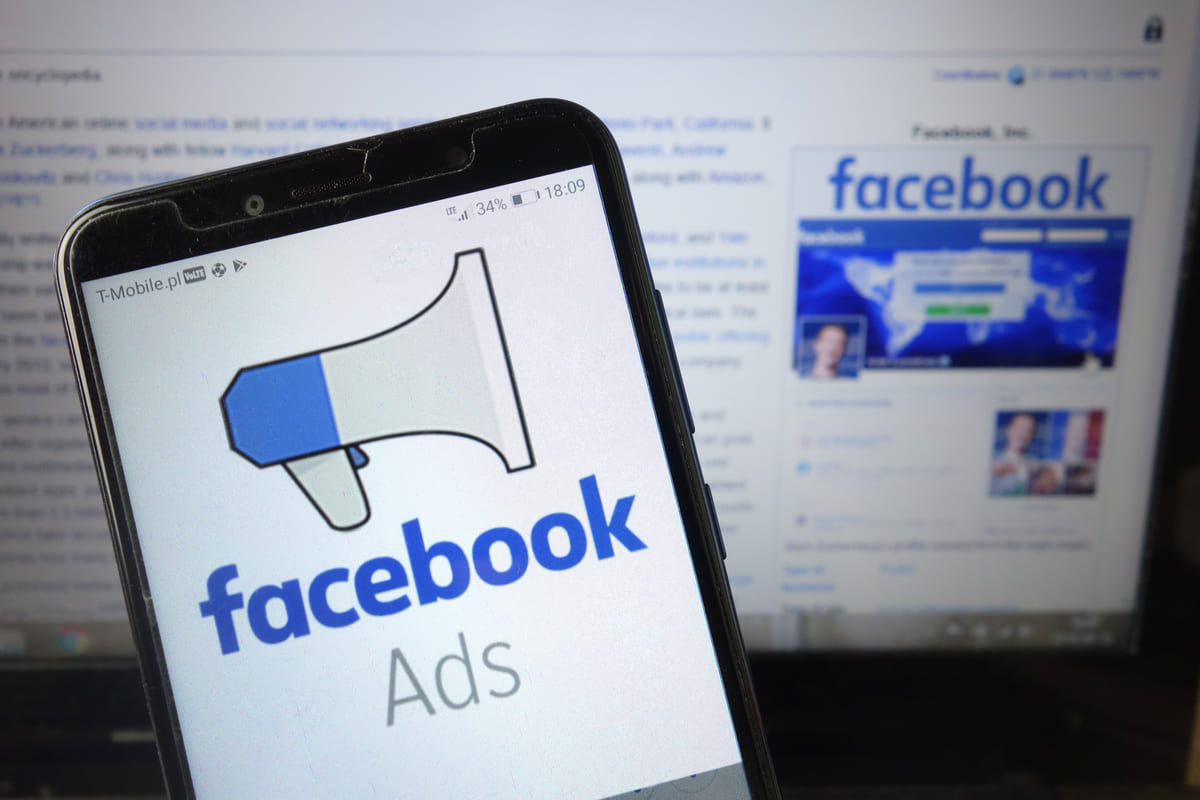 Um smartphone exibe o logotipo do Facebook Ads com um megafone estilizado, em primeiro plano, enquanto a página inicial do Facebook é visível ao fundo em um monitor. A imagem representa a utilização da plataforma de publicidade do Facebook para estratégias de marketing digital.