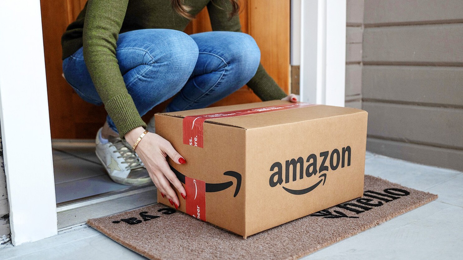 Uma pessoa se abaixa para pegar uma caixa de papelão da Amazon que foi entregue em sua porta. A imagem ilustra a etapa final da jornada de compra no e-commerce, resultado direto das estratégias de marketing digital que levaram o cliente a realizar a compra online.