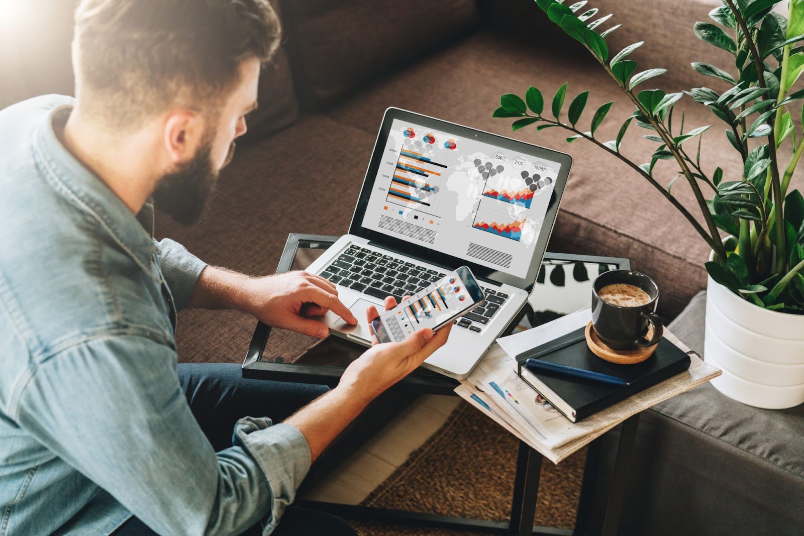 Um homem com barba, sentado em um sofá, está trabalhando em seu laptop e smartphone ao mesmo tempo. As telas de ambos os dispositivos exibem gráficos e análises de dados, demonstrando a otimização de campanhas de marketing digital. Ele segura o telefone com uma mão enquanto navega no laptop com a outra. Ao lado, há um caderno, uma caneta e uma xícara de café, sugerindo um ambiente de trabalho confortável e produtivo.