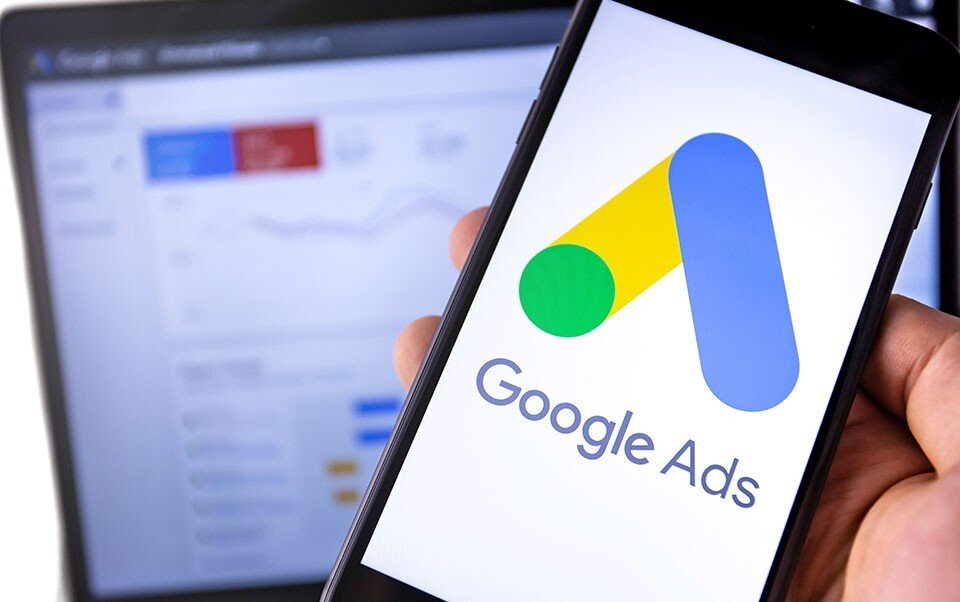 Um smartphone exibindo o logotipo do Google Ads, com um computador ao fundo mostrando um painel de análise de desempenho. A imagem ilustra a importância dos anúncios pagos no Marketing Digital, demonstrando como o Google Ads é essencial para a captação de leads e o aumento das vendas online.