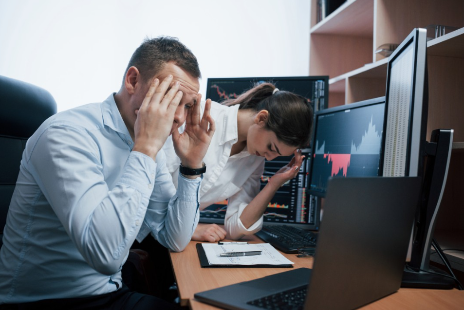 Um homem e uma mulher estão visivelmente frustrados com gráficos financeiros no fundo, sugerindo perdas. Essa imagem reflete o estresse no trabalho, algo que pode ser evitado ao investir em Infoprodutos, oferecendo um caminho mais rentável e eficiente