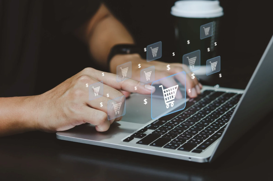 Uma pessoa está usando um laptop, com as mãos sobre o teclado. Vários ícones virtuais de carrinho de compras e símbolos de dólar flutuam acima do laptop, com um carrinho de compras maior e destacado no centro, sendo tocado por um dedo. O fundo está ligeiramente desfocado, mostrando uma xícara de café. A imagem representa o conceito de vendas online e o processo de Como Vender pela Internet.