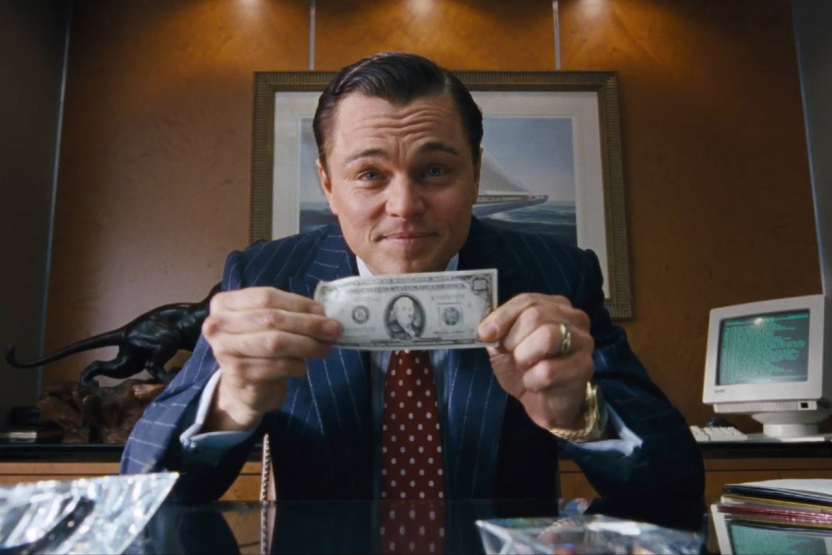 Cena do filme 'O Lobo de Wall Street', onde o personagem de Leonardo DiCaprio exibe uma nota de dólar, simbolizando a busca incessante pelo sucesso nas vendas e no mercado financeiro.