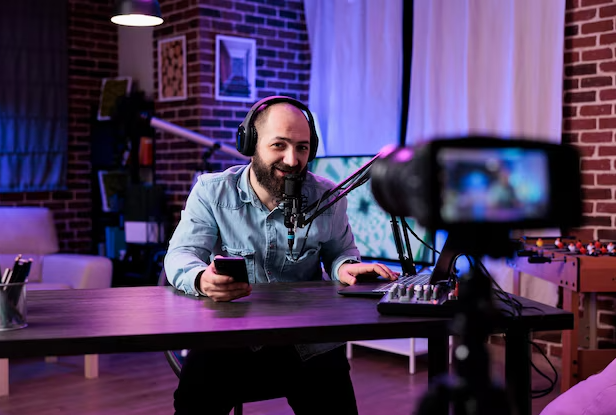 Homem gravando um curso online com um microfone e uma câmera voltada para ele, enquanto ele segura um celular. O ambiente tem iluminação moderna e aconchegante, ideal para transmissões ao vivo ou gravações de conteúdo educacional.