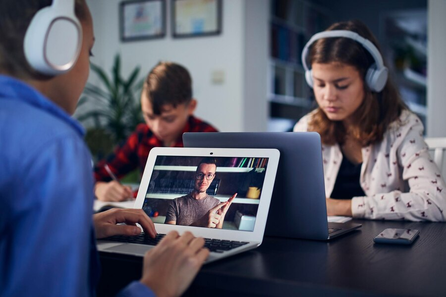 Crianças assistindo a um curso online em seus laptops, com uma aula sendo transmitida por vídeo. Elas usam fones de ouvido enquanto uma delas interage com o conteúdo na tela do laptop, onde um professor aparece explicando algo.