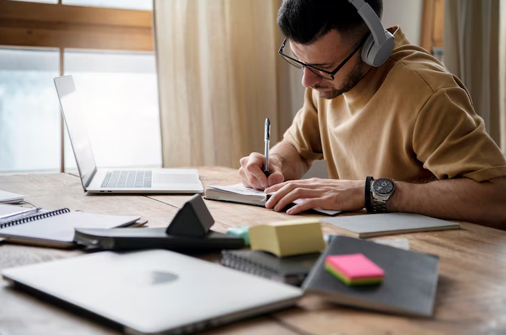Homem escrevendo em caderno enquanto estuda com fones e notebook, refletindo tendências de aprendizado remoto e autodidata.