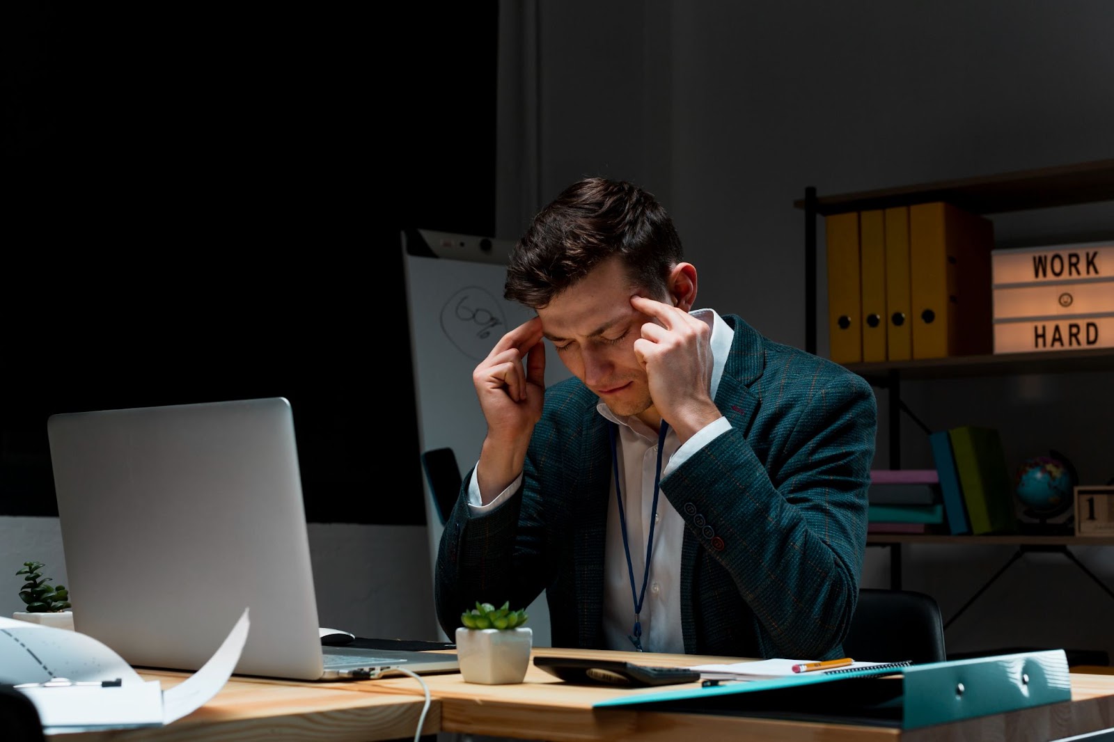 Homem sentado à mesa em um escritório, visivelmente estressado e concentrado, pressionando as têmporas com os dedos enquanto encara o notebook. Ao fundo, a luz suave de uma luminária e a frase "Work Hard" reforçam o ambiente de esforço e dedicação. A imagem retrata os desafios e a pressão enfrentados por quem busca lucrar alto, mostrando que grandes resultados exigem foco mental, resiliência e trabalho estratégico.