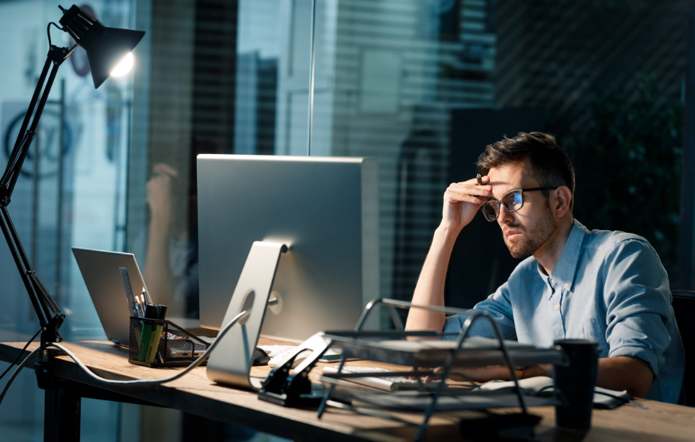 Homem com expressão pensativa em escritório moderno à noite, sentado em frente a dois computadores, refletindo sobre estratégias de como vender mais e melhorar resultados nas vendas online.