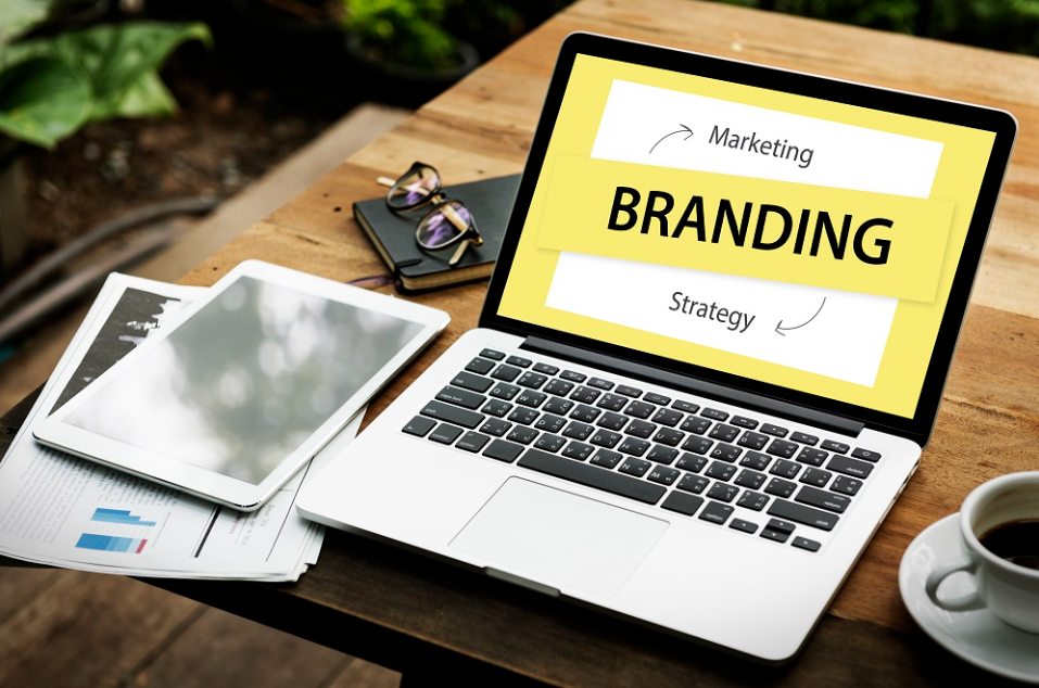 Notebook com a palavra "Branding" destacada na tela, cercado por tablet, documentos e café, representando estratégias de marketing e planejamento de branding em ambiente de trabalho criativo.