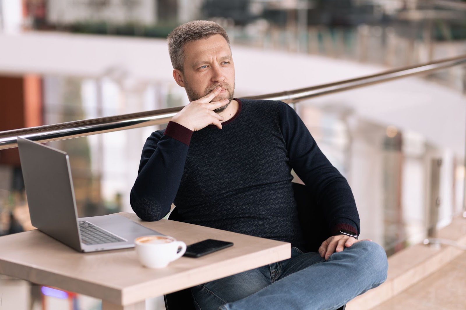 Homem pensativo sentado em uma cafeteria com notebook, celular e café sobre a mesa, refletindo sobre estratégias para seu negócio online.