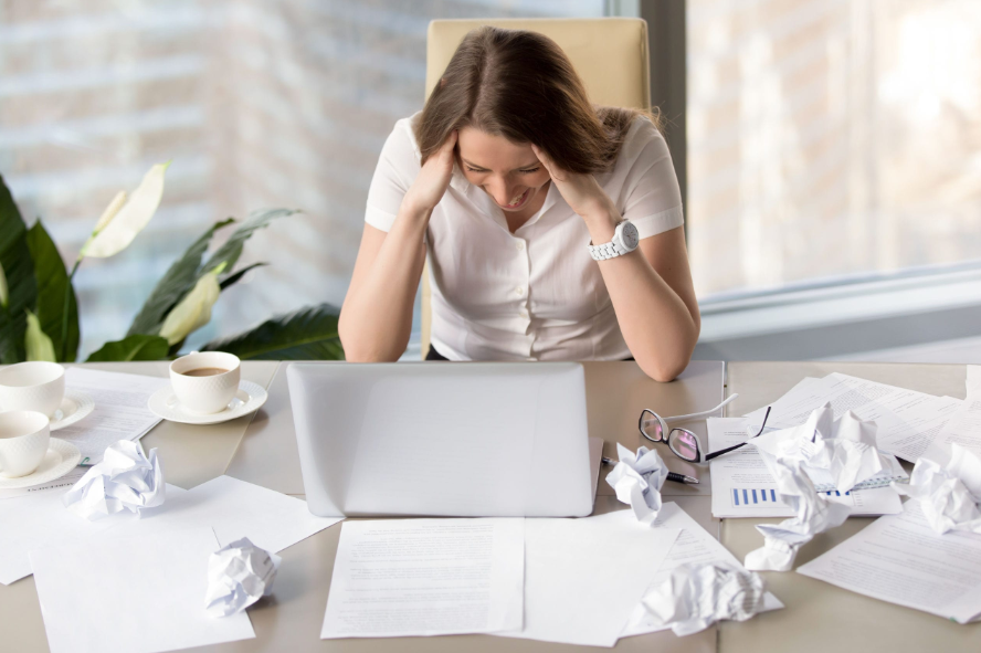 Mulher estressada diante de um laptop em meio a papéis amassados, representando os desafios de quem ainda não encontrou uma forma eficiente de trabalhar com produtos digitais.