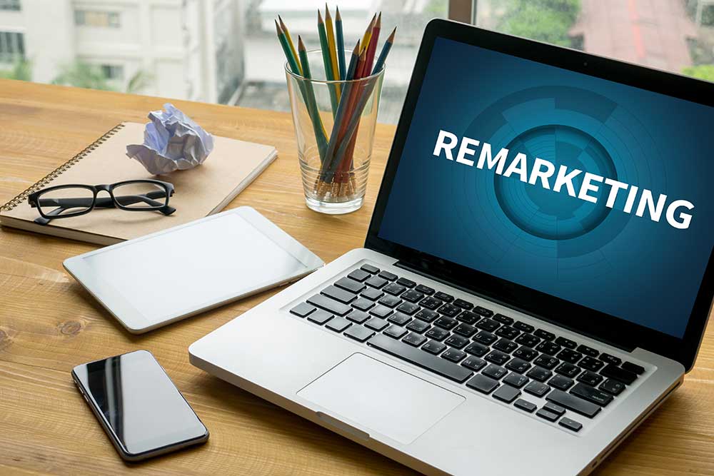 Mesa de trabalho com notebook exibindo a palavra 'Remarketing' na tela, ao lado de itens de escritório como óculos, tablet, celular, caderno e lápis, representando estratégias digitais e planejamento de remarketing.