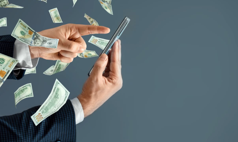 Homem interage com um smartphone enquanto notas de dinheiro flutuam ao redor, simbolizando os lucros gerados pela venda de produtos digitais através da internet.