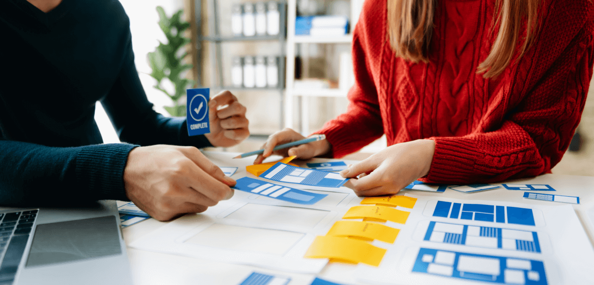 Mãos manipulando elementos de design de interface e notas em uma mesa de trabalho. Isso simboliza a busca por eficiência e o aprimoramento da experiência para guiar os 'leads' de forma eficaz.