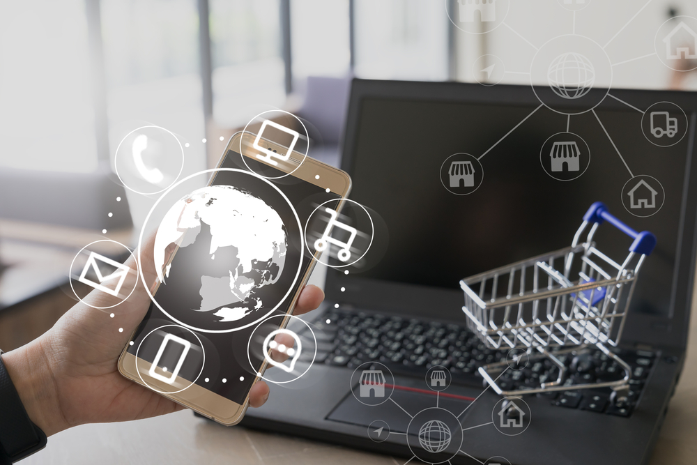 Visual de e-commerce moderno com uma mão segurando um smartphone que projeta uma rede de ícones digitais como comunicação e compras. Um laptop e um mini carrinho de supermercado estão em segundo plano. Ideal para ilustrar estratégias de Vendas no Telegram e outras plataformas de comércio online.
