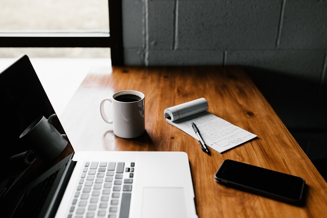 Imagem de um setup de trabalho minimalista com um notebook prateado, uma xícara de café branca, um caderno de anotações com uma caneta e um smartphone preto sobre uma superfície de madeira. Representa o espaço para idealizar e gerenciar atividades digitais, como as Vendas no Telegram.