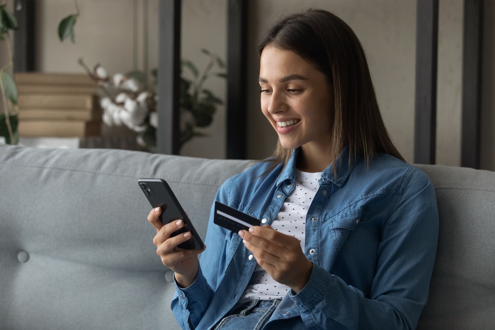 Uma mulher sorridente sentada em um sofá, segurando um smartphone na mão esquerda e um cartão de crédito preto na mão direita, digitando informações no celular. A imagem retrata uma compra online ou transação digital, relevante para processos de pagamento em Vendas no Telegram.