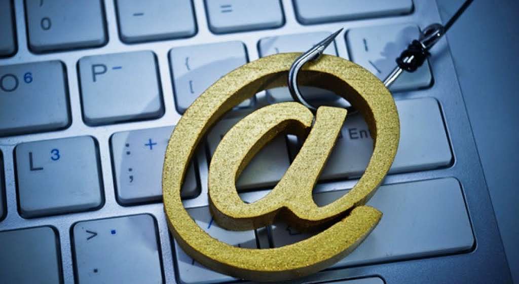 Um símbolo '@' dourado preso a um anzol sobre um teclado, talvez representando uma isca digital. De forma muito indireta, poderia-se tentar associar a ideia de uma 'isca' com um Lead Magnet, mas a imagem primariamente ilustra um ataque de phishing, não uma estratégia de marketing de conteúdo.