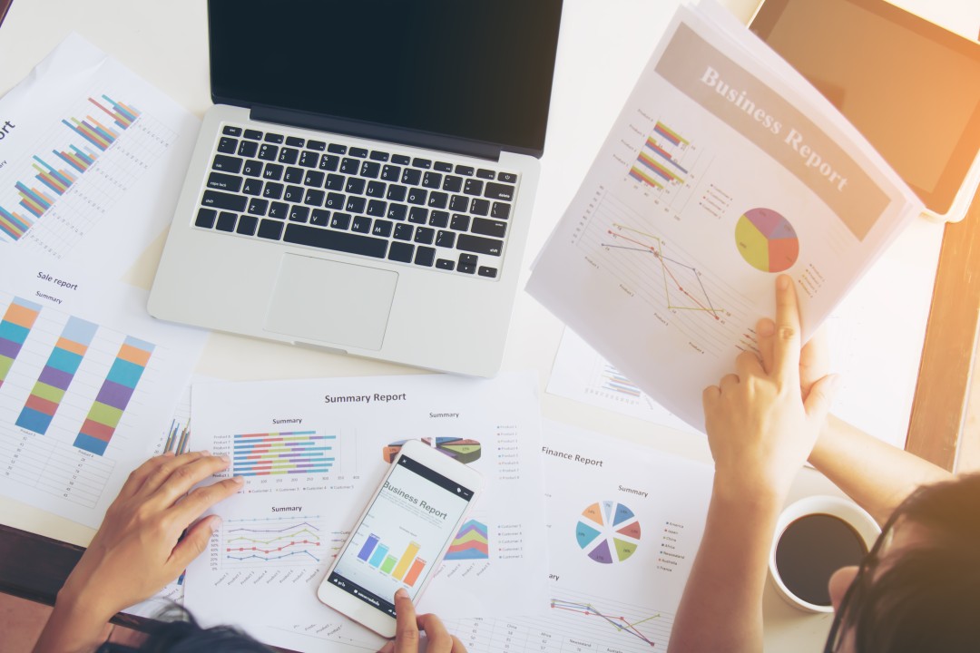 Uma mesa repleta de relatórios financeiros, gráficos de vendas e dispositivos eletrônicos (laptop, smartphone). O conteúdo detalhado e visualizado nesses documentos representa o tipo de material que pode ser usado como um Lead Magnet para atrair e educar leads.