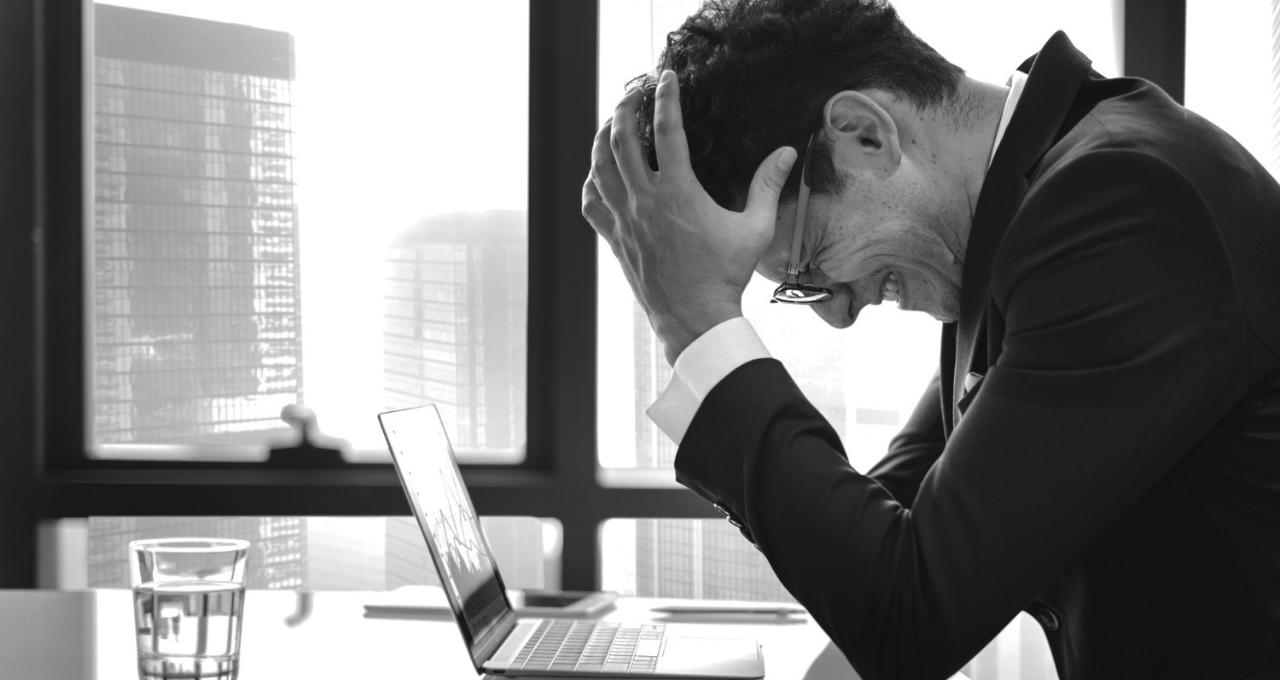 Um homem de negócios em apuros, expressando frustração com a cabeça nas mãos, enquanto um laptop está aberto em sua mesa. Talvez a 'solução' para seu problema pudesse ser um Lead Magnet para quem está passando por situação similar.